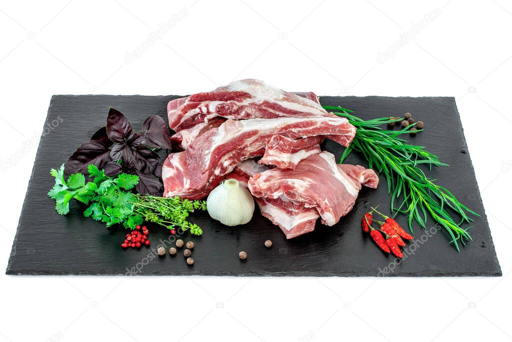 Raw pork ribs and condiments laid out on a black stone cutting board. Whole raw pork ribs with fresh herbs on dark black background. Raw pork ribs, pepper, basil, garlic and condiments