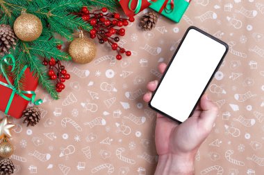 Model görüntüsü. Adamın eli Noel dekorasyonunda cep telefonu tutuyor. Cep telefonu modeli insan eliyle yapılmış. Arka planda Noel ve yeni yıl süsleri ve hediyeleri.