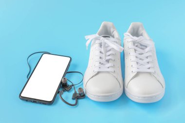 Model görüntüsü. Spor ayakkabı ve telefon. Boş ekranlı cep telefonu, mavi arka planda beyaz spor ayakkabılar. Boş Moblie akıllı telefonu, kulaklıkları ve spor ayakkabıları var. Sağlıklı yaşam biçimleri