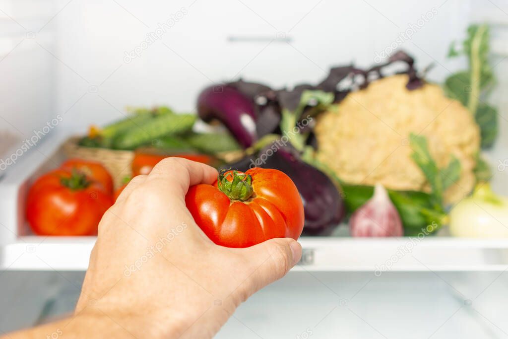 Man takes the tomato from the open refrigerator. refrigerator is full of vegetables and herbs. Healthy food. Weight loss diet concept.