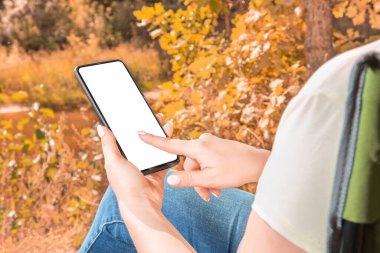 Siyah cep telefonunu elinde tutan ve gösteren bir kadının sonbahar dönemi arka planına sahip boş ekran görüntüsü. Model telefon. Sonbahar arkaplanı bulanık