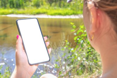 Model cep telefonu. Kadının elinde boş ekranlı bir telefon var. Gölün ve yeşilliğin arka planında. Doğanın arka planına karşı telefon modeli