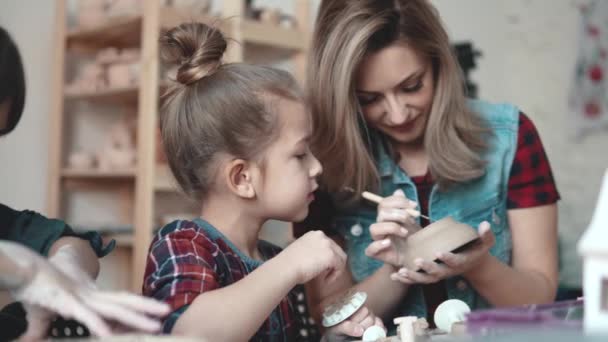 Маленькая девочка делает глиняное ремесло в керамической мастерской. Мама и ребенок проводят время вместе, выполняя творческую работу — стоковое видео