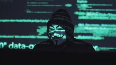 Erkek hırsız maskeli karanlık bir odada bir bilgisayarda çalışır. bilgisayar kodu yüzünde yansıtılır. hacker sistemi tatili.