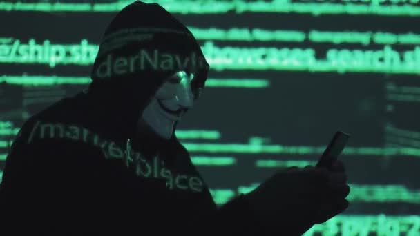 Robo de datos personales en la red. ladrón en la máscara y capucha contra el fondo del código en ejecución. anónimo utiliza smartphone para romper el sistema — Vídeo de stock
