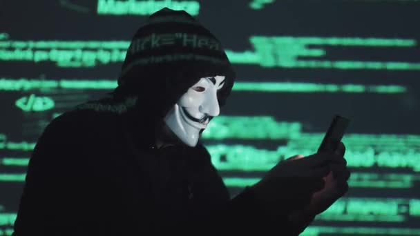 Diebstahl personenbezogener Daten im Netzwerk. Räuber in Maske und Kapuze vor dem Hintergrund des laufenden Codes. Anonymous nutzt Smartphone, um System zu knacken — Stockvideo