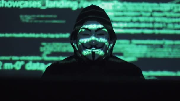 Robo de datos personales en la red. ladrón en la máscara y capucha contra el fondo del código en ejecución. anónimo utiliza una computadora para entrar en el sistema — Vídeo de stock