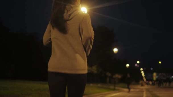 Corre por la ciudad por la noche. Una joven deportista corriendo a lo largo del terraplén a la luz de farolas. vista posterior — Vídeos de Stock