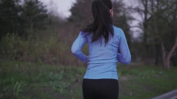 Girl trains outdoors. jogging. A young girl in blue sports clothes is running along the evening park. back view. female legs in sneakers close-up — Stock Video