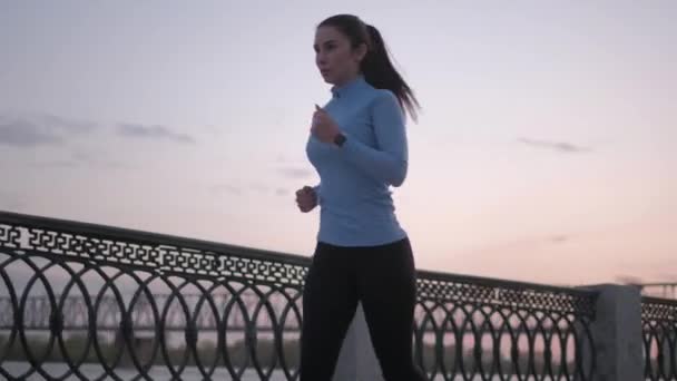 Menina desportiva em um relógio inteligente corre ao longo do dique ao pôr do sol. retrato de uma esportista fazendo um esporte na natureza — Vídeo de Stock