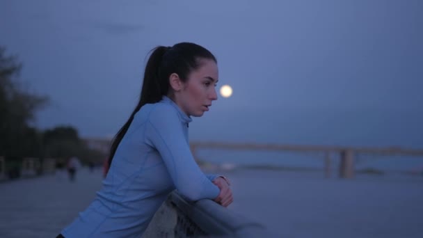 Fille en vêtements de sport repose ses coudes sur la clôture sur le remblai. courir à travers la ville — Video