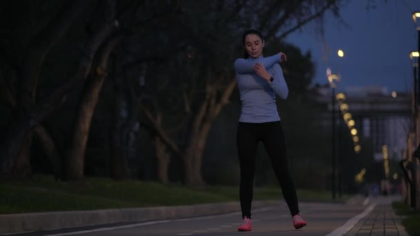 Alongamento antes de iniciar o treino. jovem menina desportiva aquece antes de correr no parque da noite — Vídeo de Stock