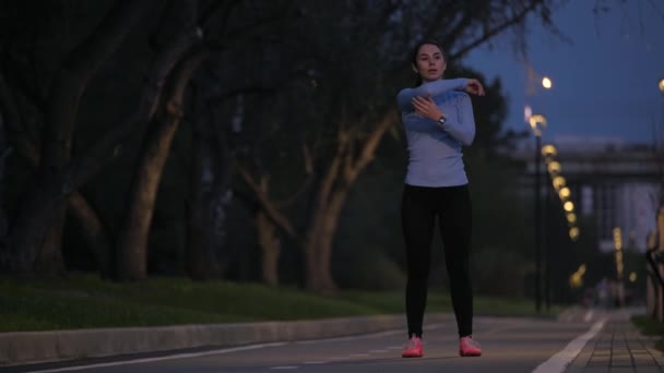 Растяжение перед началом тренировки. Молодая спортивная девушка разогревается перед пробежкой в вечернем парке — стоковое видео