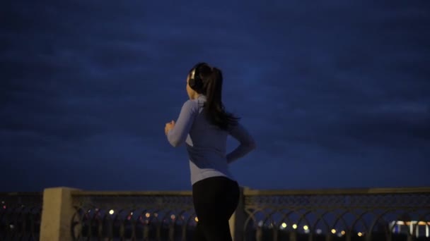 Menina ouve música com fones de ouvido enquanto Jogging na cidade à noite. desportista em sportswear no fundo da cidade à noite. vista para trás. menina alegremente levanta as mãos no final do treinamento — Vídeo de Stock