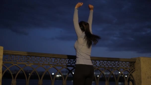 Chica atleta con una hermosa figura corre a lo largo del terraplén por la noche. deportista termina el entrenamiento y felizmente levanta las manos. una mujer joven disfruta de sus logros deportivos y éxitos — Vídeos de Stock