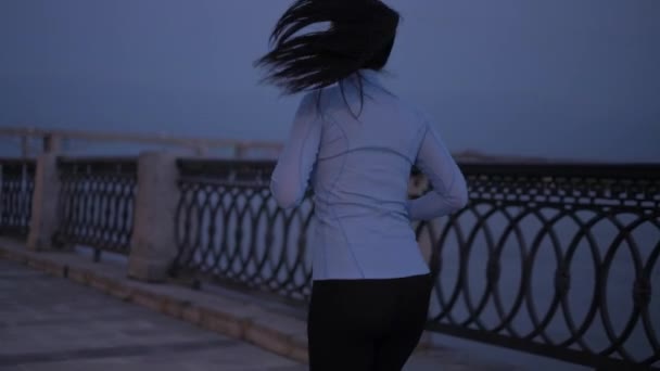 Une jeune fille en vêtements de sport Jogging sur le front de mer au clair de lune. l'athlète est engagé dans des sports sur le fond des lumières de la ville la nuit. au ralenti — Video