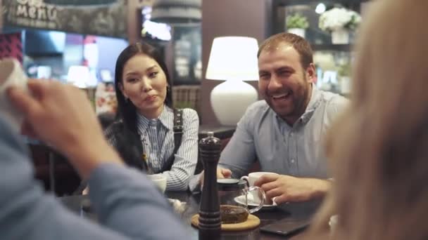 Друзья проводят время вместе за ужином в ресторане. молодой человек и девушка разговаривают с друзьями и улыбаются — стоковое видео
