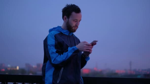 若い男は、夕暮れ時、携帯電話を使用します。男は彼のスマート フォンと笑顔のメッセージを送信します。 — ストック動画