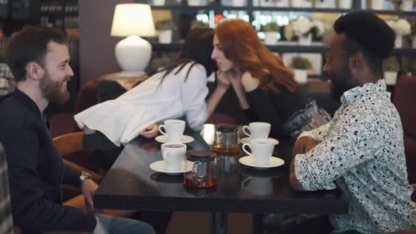 Multietnické společnosti přátel v kavárně nebo restauraci. přátelé poraďte a bavit se společně. dvě holky a dva kluci u stolu v útulné kavárně. holky drbou na pozadí — Stock video