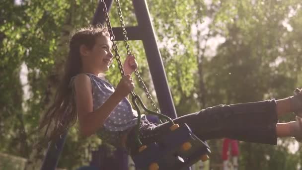 Concept of a happy and carefree childhood. child swings on a swing and laughs enthusiastically. little girl on a swing in the sunlight. slow motion — Stock Video