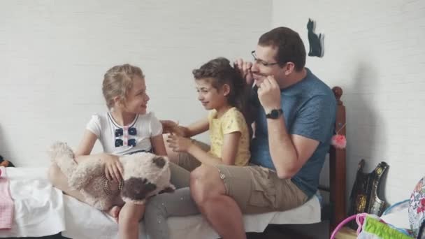 Молодий батько грає зі своїми маленькими дітьми. тато і дві дочки сидять в дитячій кімнаті на ліжку — стокове відео