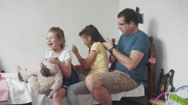 Ein junger Vater flechtet seinen Töchtern Zöpfe. Familienporträt. ältere Schwester flechtet Zöpfe jüngere Schwester — Stockvideo