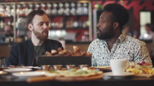 Dva mladí muži jíst steak v restauraci. dva přátelé sedí u stolu v útulné restauraci a bavte se mluví — Stock video