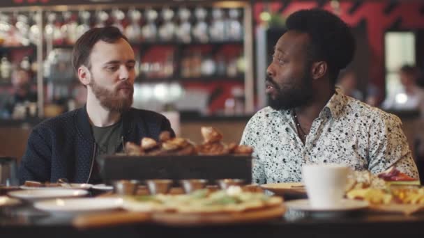 Dos jóvenes comen un bistec en un restaurante. dos amigos se sientan en una mesa en un acogedor restaurante y se divierten hablando — Vídeos de Stock