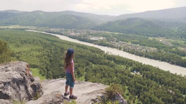 Un piccolo viaggiatore ammira la vista in piedi su una scogliera su una roccia. ragazza turista in montagna — Video Stock