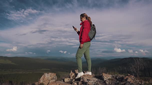 Jeune voyageuse avec sac à dos et chapeau fait un selfie sur téléphone portable debout sur le bord de la falaise dans les montagnes — Video