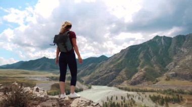 Bir gezgin bir sırt çantası ile dağlarda bir uçurumun kenarında duruyor ve ellerini yukarı yükseltir. Arkadan görünüm