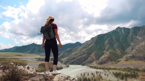 Un viajero con una mochila está de pie en el borde de un acantilado en las montañas y levanta las manos. vista posterior — Vídeo de stock