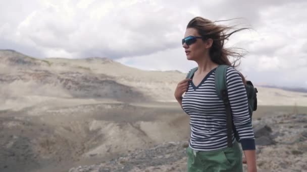 Turista chica va en un terreno montañoso. retrato del viajero. mujer excursionista con mochila, montañas en el fondo . — Vídeo de stock