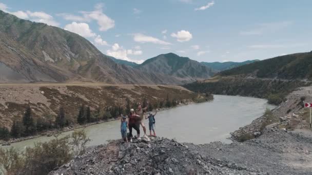 Antény. cestující s batohy usmívat se a mávat rukama ve fotoaparátu. rodina turistů v horách nedaleko horské řeky — Stock video