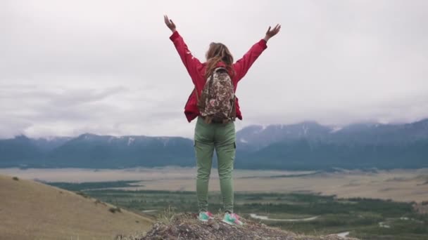 Touristenmädchen mit Rucksack steht am Rand der Klippe und genießt den weiten Blick ins Tal. der Reisende hebt die Hände und jubelt nach oben über das Erreichen des Ziels. — Stockvideo