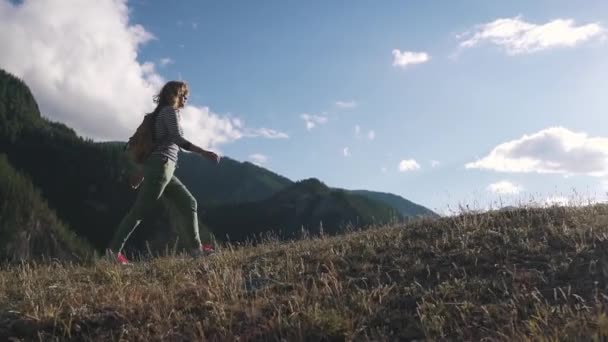 Chica viajero en el fondo de las montañas y las nubes. Un turista con una mochila se levanta cuesta arriba . — Vídeos de Stock