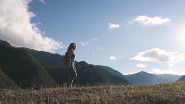 Ένας ταξιδιώτης με ένα σακίδιο περιπάτους κατά μήκος της κορυφής του λόφου στο παρασκήνιο των βουνών. κορίτσι τουριστικά στα βουνά — Αρχείο Βίντεο