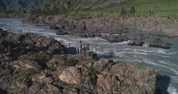 Aerea. famiglia di turisti. i viaggiatori si trovano sul bordo di una scogliera sopra un fiume di montagna. genitori con bambini in escursione. vista posteriore — Video Stock