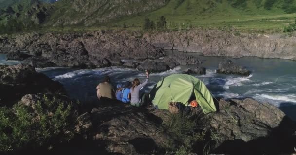 Aéreo. Uma família de viajantes com crianças se senta perto de uma tenda e desfruta de uma vista do rio da montanha e das montanhas . — Vídeo de Stock