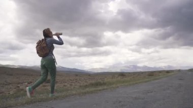 Gezgin içecekler su üstünde belgili tanımlık gitmek. Kız turistik bir sırt çantası ve dağların fon karşı giyen güneş gözlüğü