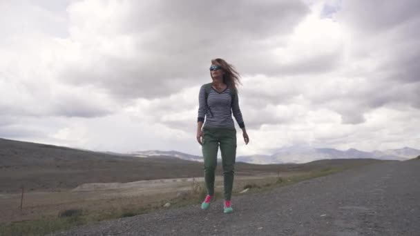 Путешественник проходит по горной местности и оборачивается. девушка туристка с рюкзаком и в солнечных очках на фоне гор — стоковое видео