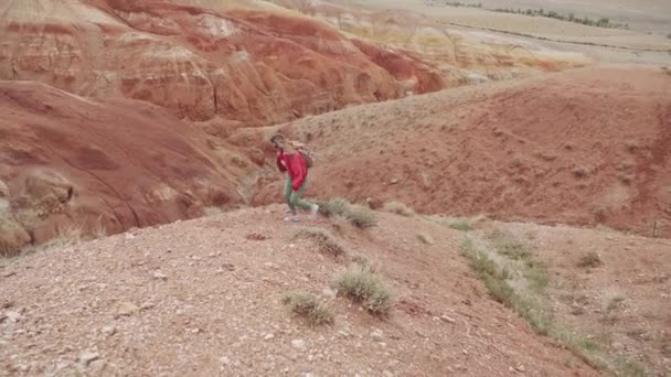 旅行者登上陡峭的山坡。一个带着背包的旅游女孩戴着太阳镜沿着山地的地形行走. — 图库视频影像