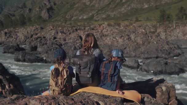 En ung resenär mamma sitter på kanten av en klippa nära en mountain river med sina barn. familj av turister. bakifrån — Stockvideo