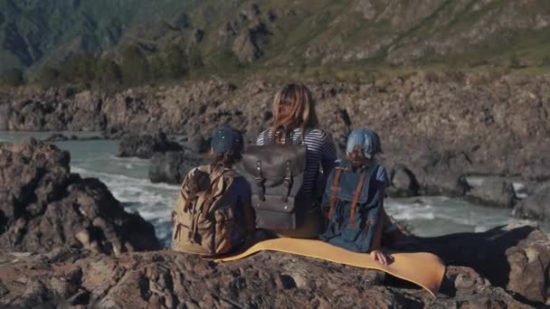 Una joven madre viajera se sienta en el borde de un acantilado cerca de un río de montaña con sus hijos. familia de turistas. vista posterior — Vídeos de Stock