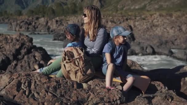 Eine junge reisende Mutter sitzt mit ihren Kindern am Rande einer Klippe in der Nähe eines Gebirgsflusses. Touristenfamilie. — Stockvideo