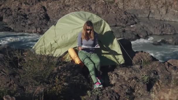 Flicka studerar kartan sitter av tältet på kanten av en klippa nära en mountain river — Stockvideo