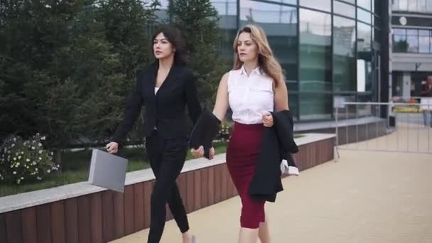 Das Mädchen im Businessanzug kommt mit einem Kollegen in Rock und Bluse. Geschäftsfrauen laufen die Straße entlang gegen ein gläsernes modernes Gebäude — Stockvideo