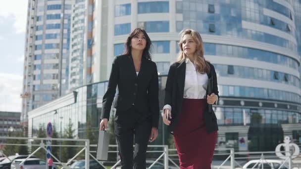 Dos mujeres atractivas jóvenes en trajes formales van contra el fondo de un moderno centro de negocios — Vídeos de Stock
