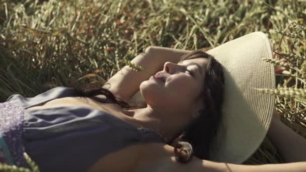 Una chica con una paja en la boca se encuentra en el campo y disfruta de un cálido día de verano. Joven mujer hermosa en un sombrero de paja y ropa rústica. concepto de recreación veraniega y unidad con la naturaleza — Vídeos de Stock