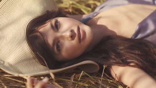 Chica encantadora en un sombrero de paja se encuentra en el campo mirando y sonriendo a la cámara. Retrato de cerca de una hermosa joven — Vídeos de Stock